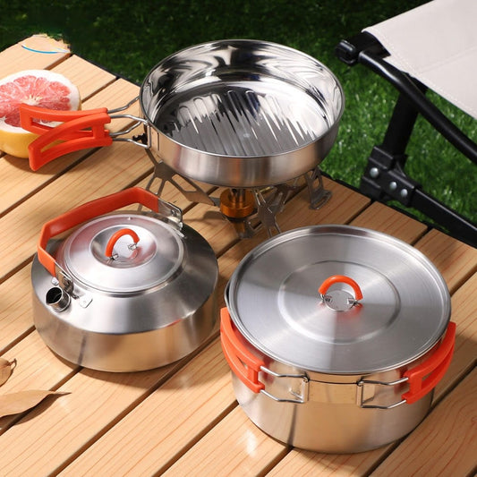 Set of stainless steel cookware with orange handles on a wooden surface outdoors.